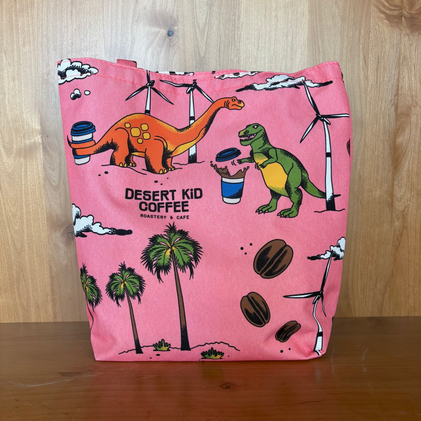 Pink tote bag with dinosaur design and 'Desert Kid Coffee' branding on a wooden surface.