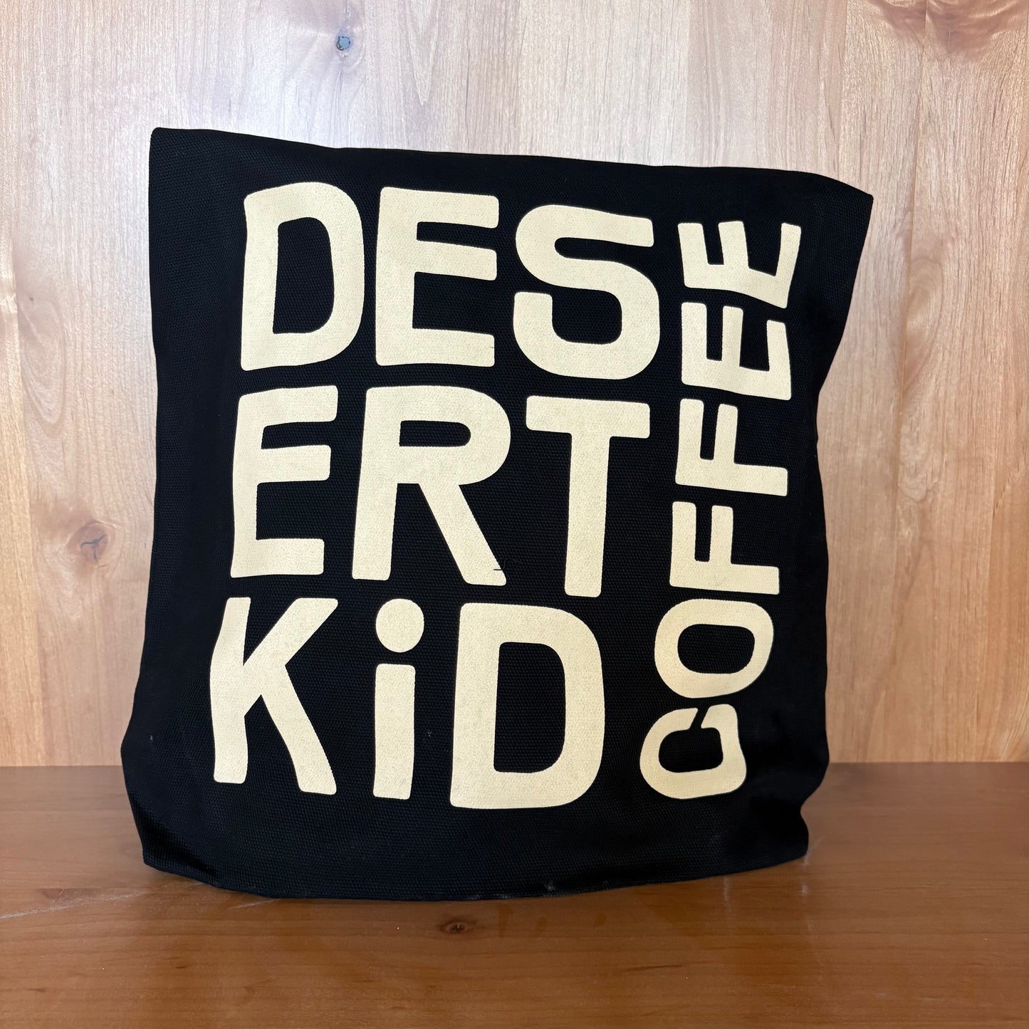 Black pillow with white text 'DESERT KID COFFEE' on a wooden surface