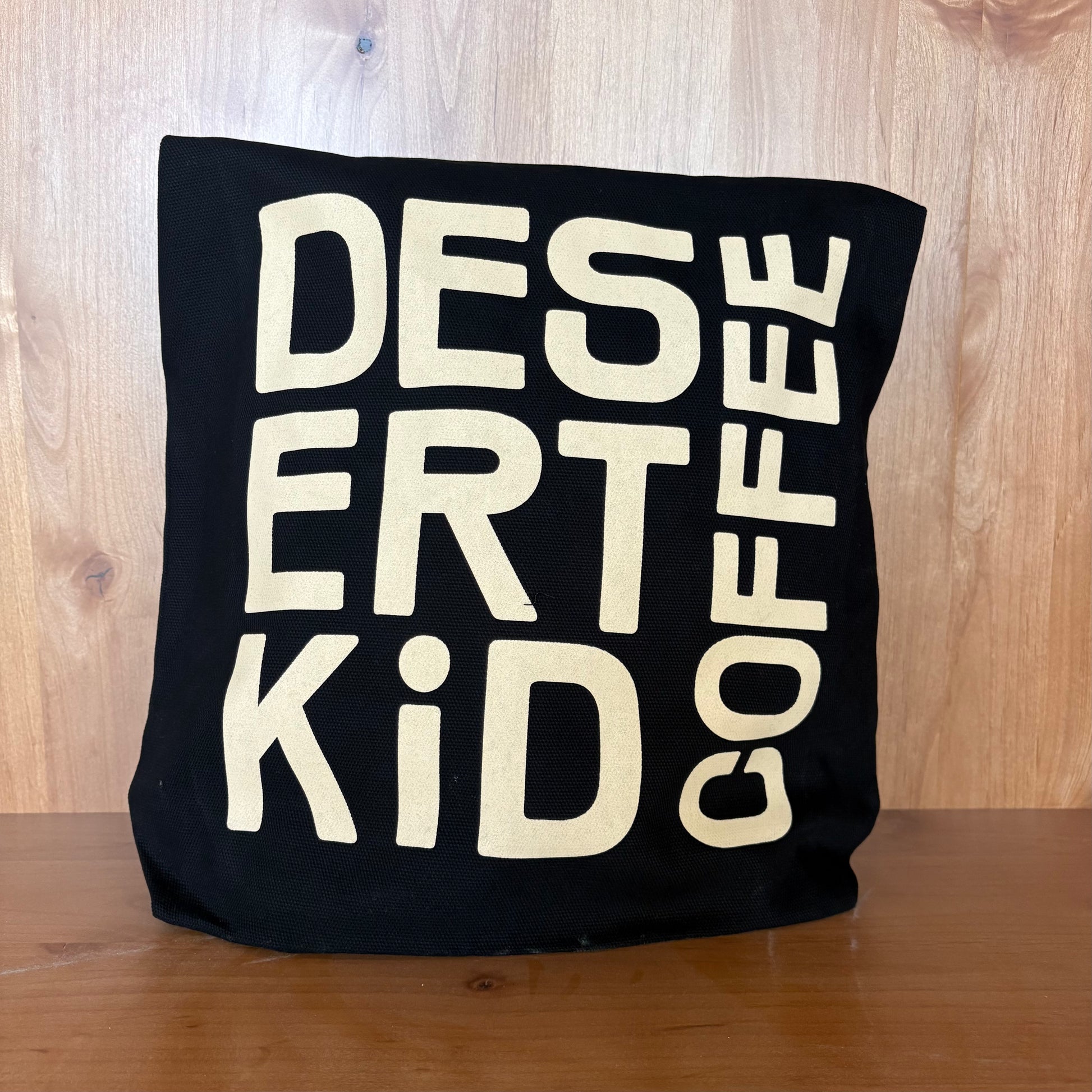 Black pillow with white text 'DESERT KID COFFEE' on a wooden surface